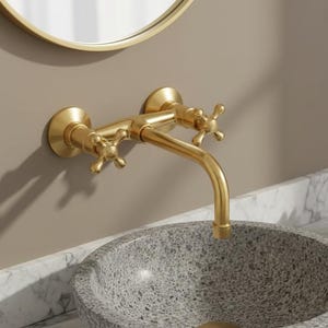 Peut inclure: Un robinet mural doré avec des poignées en forme de croix est monté au-dessus d'un évier gris moucheté. Un miroir rond doré est visible en arrière-plan. Une petite plante succulente dans un pot blanc et des serviettes blanches pliées sont sur le comptoir en marbre.