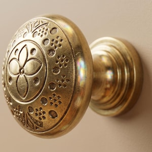 Puede incluir: Primer plano de un pomo de armario de latón con un diseño floral ornamentado. El pomo es redondo con un patrón detallado de flores y puntos. El herraje está montado sobre una superficie de color neutro.
