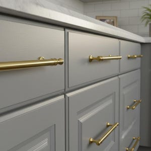 Peut inclure: Gros plan sur une armoire de cuisine avec des portes et des tiroirs gris. Les armoires sont équipées de poignées de barre dorées. Le comptoir est blanc et une petite plante est visible en arrière-plan.