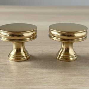 May include: Two gold-colored cabinet knobs with a round top and a decorative base. The knobs are made of metal and have a shiny finish. They are designed for use on drawers or cabinets.