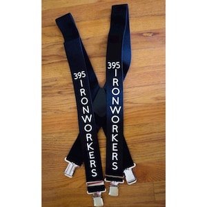May include: Black suspenders with the text "395 IRONWORKERS" in white. The suspenders have silver-colored metal clips and adjusters. The suspenders are lying on a wooden surface.