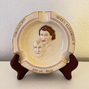 Puede incluir: Cenicero crema con un retrato de la reina Isabel II y el príncipe Felipe. El cenicero tiene las palabras "CORONATION" y "QUEEN ELIZABETH II" en azul y rojo. También se ve la fecha "2 de junio de 1953".