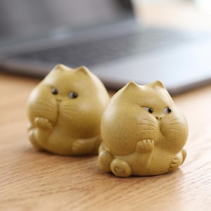 May include: Two small, yellow cat figurines with a rounded shape and a slightly grumpy expression. The figurines are sitting on a wooden surface. The cat figurines have small paws and a heart-shaped detail on the bottom.