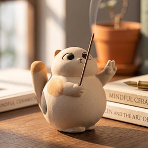 May include: A ceramic cat figurine in a light beige color, holding an incense stick. The cat has brown spots and a raised paw. The figurine is on a wooden surface, with books and a potted plant in the background.