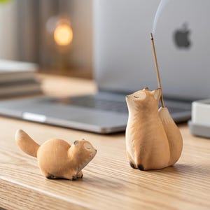 May include: Two light brown fox figurines made of matte material. One fox is in a sitting position with a thin incense stick inserted into its back, emitting a trail of smoke. The other fox is positioned horizontally in a lying down pose.