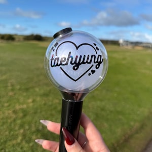 Puede incluir: Un bastón de luz negro y plateado con una parte superior esférica transparente. La parte superior presenta un diseño de corazón con el nombre "taehyung" en cursiva. El mango tiene las palabras "MAP OF" impresas. El fondo es un campo verde y cielo azul.