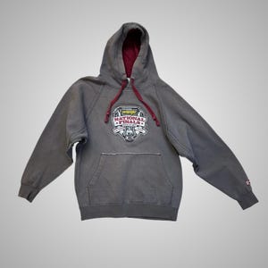 Op de afbeelding: Grijze Wrangler National Finals Rodeo hoodie met een kangoeroezak en een capuchon met trekkoord. De voering van de capuchon en de trekkoorden zijn bordeauxrood. De voorkant heeft een logo met de tekst "Wrangler National Finals Rodeo".