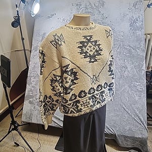 May include: Cream-colored sweater with a geometric black pattern. The sweater is displayed on a mannequin, showcasing its design and texture. The pattern includes shapes and lines, creating a detailed and visually appealing design. The sweater is paired with a black skirt.