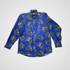 Puede incluir: Camisa azul abotonada con un patrón repetido de vehículos marrones y verdes. La camisa de manga larga tiene cuello y un bolsillo en el pecho. La tela tiene un aspecto ligeramente descolorido.