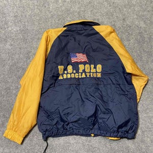 May include: A navy blue and yellow windbreaker jacket featuring the text U.S. POLO ASSOCIATION in gold lettering and an embroidered American flag on the back. The jacket has long yellow sleeves, a collar, and an elasticized hem with a drawstring.