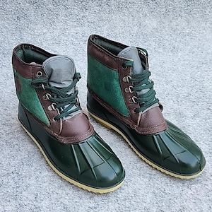 Könnte beinhalten: Ein Paar grün-braune Duck Boots. Die Stiefel haben eine grüne Gummibasis, braune Lederakzente und grüne Schnürsenkel. Der obere Teil des Stiefels ist aus grünem Wildleder und braunem Leder. Die Stiefel sind für den Außenbereich konzipiert.