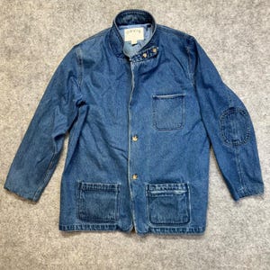 May include: A blue denim jacket with a stand-up collar, long sleeves, and elbow patches. The jacket features a button-front closure, one chest pocket, and two lower front pockets. A white label with the text ORVIS is visible at the inner neckline.