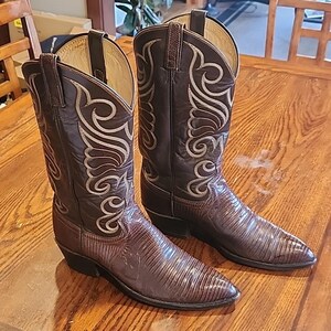 Op de afbeelding: Een paar donkerbruine leren cowboylaarzen met een textuur van hagedissenhuid op de voet en een hoge schacht met gedetailleerd crèmekleurig stiksel. De laarzen zijn voorzien van een puntige neus, een hak en treklussen aan de bovenzijde.
