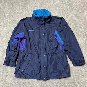 May include: Navy blue Columbia jacket with a high collar and a drawstring waist. The jacket features blue and purple accents on the sleeves and collar. It has a button-up front and two front pockets. The Columbia logo is visible on the chest.