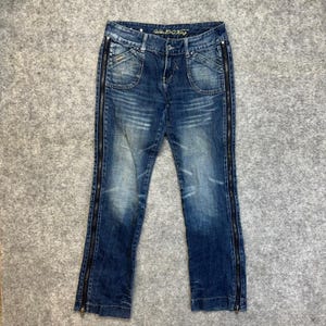 Op de afbeelding: Blauwe spijkerbroek van denim met een vervaagde wassing, een knoop- en ritssluiting en opvallende zwarte ritsen die verticaal langs de buitenste naden van elke broekspijp lopen. Het label aan de binnenzijde toont de tekst Golden E+O King in goud.
