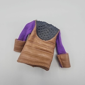 Puede incluir: Un chaleco marrón y negro de figura de acción con mangas moradas. El chaleco tiene líneas horizontales y un cuello negro con tachuelas. Las mangas tienen puños marrones.