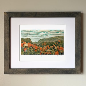 May include: Framed watercolour landscape art print showing an autumnal scene. The artwork depicts a forest with orange and green trees under a cloudy sky. The print is matted and set in a dark grey wooden frame.
