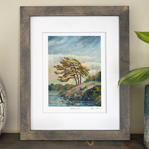 May include: Framed art print of a landscape scene. The artwork depicts a windswept tree on a rocky shoreline, with a body of water in the foreground and a cloudy sky above. The frame is a weathered grey colour.