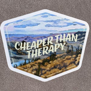 May include: A sticker with a landscape painting and the text "CHEAPER THAN THERAPY" in white. The artwork depicts a mountain range, a river, and a cloudy sky, all within a white-bordered, hexagonal shape.