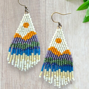 May include: A pair of handcrafted beaded earrings with a triangular design. The earrings feature a white top with an orange sunburst pattern, followed by layers of purple, green, and blue beads. The bottom has dangling white and yellow fringe.