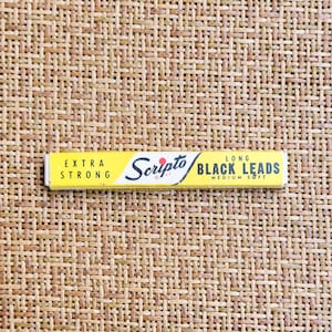 May include: A rectangular yellow box of Scripto long black leads. The packaging features the text EXTRA STRONG on the left, the Scripto logo in the center, and LONG BLACK LEADS MEDIUM SOFT printed in black on the right side of the yellow box.