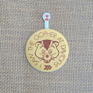 May include: A vintage button with the text "I SAW THE GOPHER AT DAYTON'S" encircling a cartoon gopher face. The button is light yellow with brown text and a brown gopher. It has a small, white tab at the top.