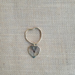 May include: A gold-colored heart-shaped pendant with a spiral design, suspended from a matching ring. The pendant is centered, and the background is a textured, neutral fabric.