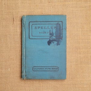 May include: A vintage blue hardcover book titled Speller Book Two, California State Series. The front cover features dark blue text and a rectangular border, with visible wear and a dark ink stain near the center of the title area.