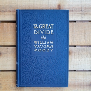 May include: A hardcover book titled "The Great Divide" by William Vaughan Moody. The book has a dark blue cover with embossed floral patterns and gold lettering. The book is closed and resting on a wooden surface.