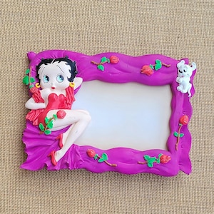 May include: A rectangular purple picture frame featuring a sculpted Betty Boop figure in a red dress on the left side, a small white dog on the top right, and several red rose accents with green stems and leaves embossed along the wavy border.
