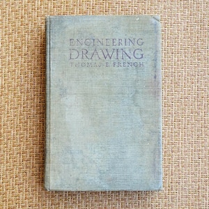 May include: A vintage hardcover book titled Engineering Drawing by Thomas E. French. The book features a faded, light gray-blue cloth cover with dark text printed on the front. The surface of the cover shows signs of wear and age-related discoloration.