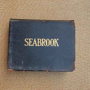 May include: A rectangular, black, hard-cover book with the word SEABROOK embossed in gold lettering on the front cover. The edges of the cover show signs of wear with some brown material visible along the corners and the spine of the book.