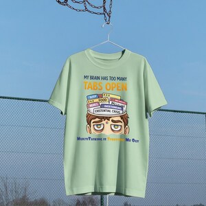 Op de afbeelding: Een lichtgroen t-shirt met de tekst "MY BRAIN HAS TOO MANY TABS OPEN" in geel en wit. Het shirt heeft een cartoonillustratie van een persoon met tabs met de labels "SLEEP", "FOOD" en "EXISTENTIAL CRISIS" op hun hoofd. De tekst "MultiTasking It's Freaking Me Out" staat eronder.