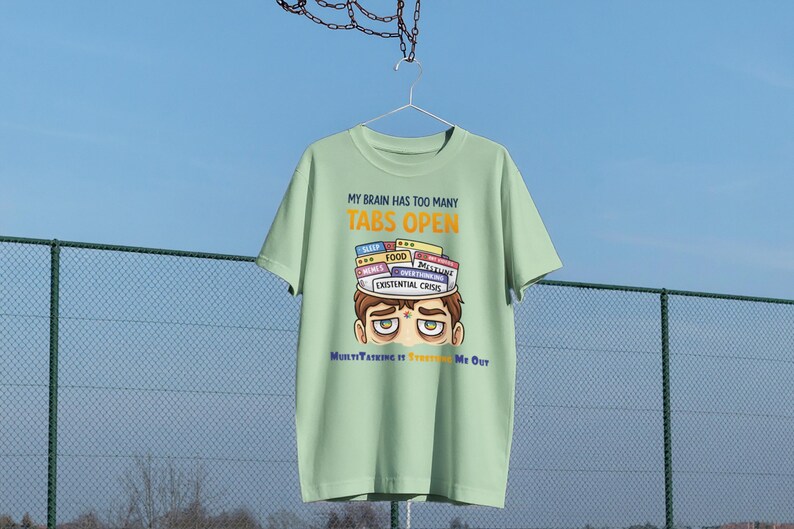 Op de afbeelding: Een lichtgroen t-shirt met de tekst "MY BRAIN HAS TOO MANY TABS OPEN" in geel en wit. Het shirt heeft een cartoonillustratie van een persoon met tabs met de labels "SLEEP", "FOOD" en "EXISTENTIAL CRISIS" op hun hoofd. De tekst "MultiTasking It's Freaking Me Out" staat eronder.