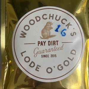 May include: A gold-colored, metallic pouch with a circular label. The label features a cartoon groundhog, the text "WOODCHUCK'S", "PAY DIRT Guaranteed SINCE 2015", and "LODE O' GOLD". The number "16" is written in blue ink.