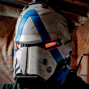 May include: A futuristic helmet with a weathered, off-white finish and blue accents. The helmet features a prominent blue stripe design and visible rivets. The eye openings glow with an orange light, suggesting an integrated display or lighting system. The overall design evokes a sci-fi or cosplay aesthetic.