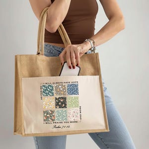 May include: A beige burlap tote bag with a cream-colored front pocket featuring a floral pattern and the text "I WILL ALWAYS HAVE HOPE" and "I WILL PRAISE YOU MORE" with a Bible verse reference. The bag has sturdy handles and is held by a person.