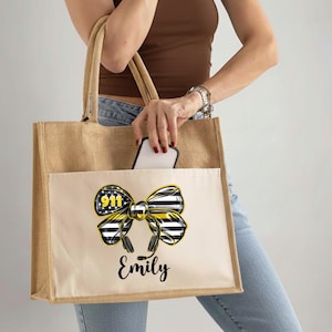 May include: A beige tote bag with a front pocket featuring a black, white, and yellow bow design with the text "911" and "Emily". The bag has a natural jute texture and sturdy handles.
