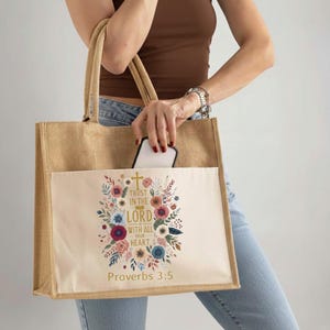 May include: A beige burlap tote bag with a cream-colored front pocket. The pocket features a floral design with the text "Trust in the Lord with all your heart" and "Proverbs 3:5". The bag has two beige handles.