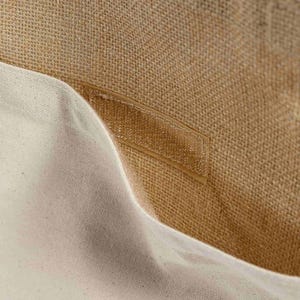 May include: Close-up of a beige burlap tote bag interior with a cream-colored canvas pocket. A clear, rectangular label is sewn onto the burlap. The bag's texture is visible, highlighting the natural fibers and construction.