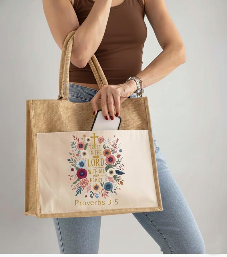 May include: A beige burlap tote bag with a cream-colored front pocket. The pocket features a floral design with the text "Trust in the Lord with all your heart" and "Proverbs 3:5". The bag has two beige handles.