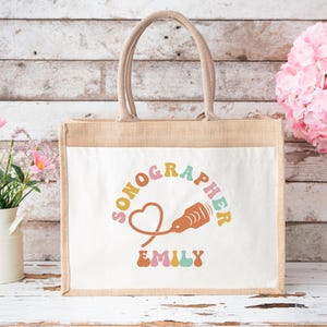 May include: A beige burlap tote bag with a white front panel. The panel features the word "SONOGRAPHER" in a retro font, along with a heart and an ultrasound image. The name "EMILY" is below the image. The bag has a jute handle.