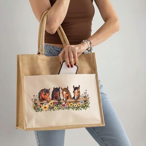 May include: A beige tote bag with a pocket featuring a design of four horse heads peering over a wooden fence with colorful flowers. The bag has sturdy handles and is held by a person.