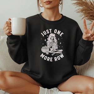 May include: Black crewneck sweatshirt with the text "JUST ONE MORE ROW" and a graphic of a skeleton knitting. The skeleton is seated with a basket of yarn and knitting needles. The sweatshirt is a solid black color.