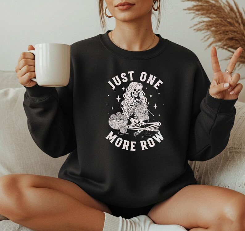 May include: Black crewneck sweatshirt with the text "JUST ONE MORE ROW" and a graphic of a skeleton knitting. The skeleton is seated with a basket of yarn and knitting needles. The sweatshirt is a solid black color.