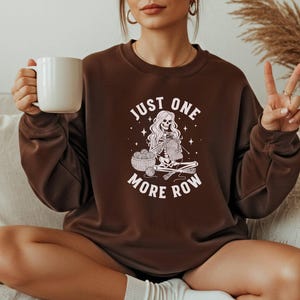May include: A brown sweatshirt with the text "JUST ONE MORE ROW" and a graphic of a skeleton knitting. The skeleton has long hair and is sitting with a basket of yarn and knitting needles. The person in the image is holding a white mug.