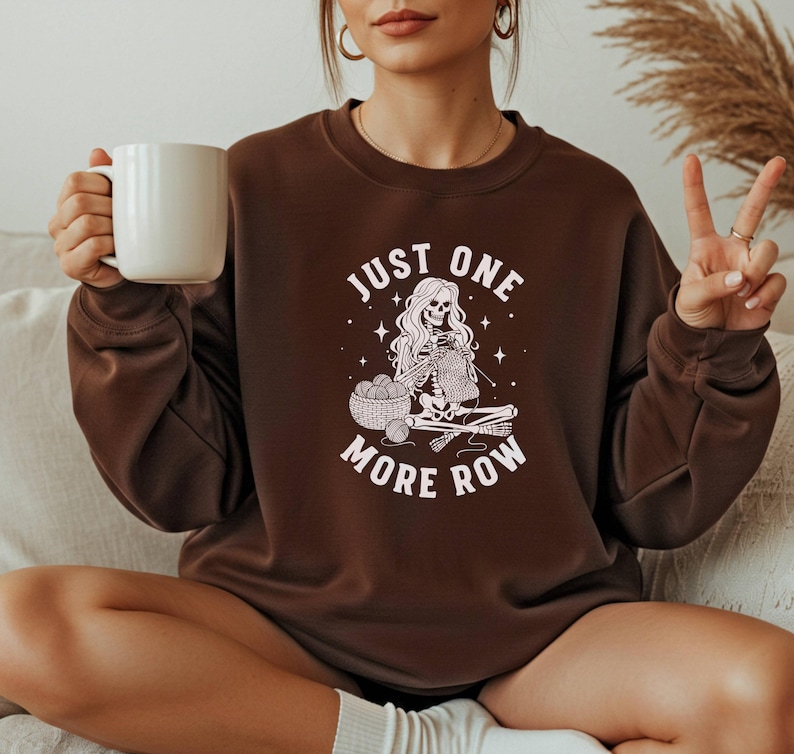 May include: A brown sweatshirt with the text "JUST ONE MORE ROW" and a graphic of a skeleton knitting. The skeleton has long hair and is sitting with a basket of yarn and knitting needles. The person in the image is holding a white mug.