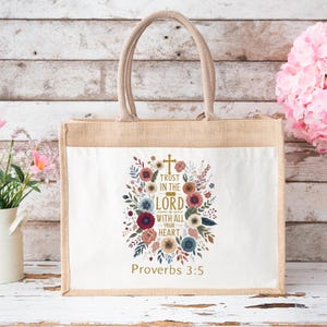 May include: A beige tote bag with a white front panel featuring a floral design and the text "Trust in the Lord with all your heart" and "Proverbs 3:5". The bag has a jute body and a beige handle.