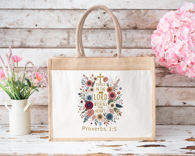 May include: A beige tote bag with a white front panel featuring a floral design and the text "Trust in the Lord with all your heart" and "Proverbs 3:5". The bag has a jute body and a beige handle.