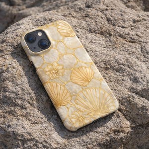 Puede incluir: Funda de teléfono crema con diseño de conchas marinas y contornos dorados. La funda, adornada con motivos de conchas y flores, descansa sobre una roca gris rugosa.
